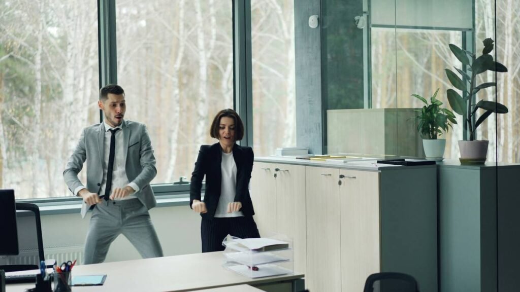 Joyful couple of colleagues is dancing in office having fun together moving arms and body relaxing after work. Millennials, carefree youth lifestyle and joy concept.