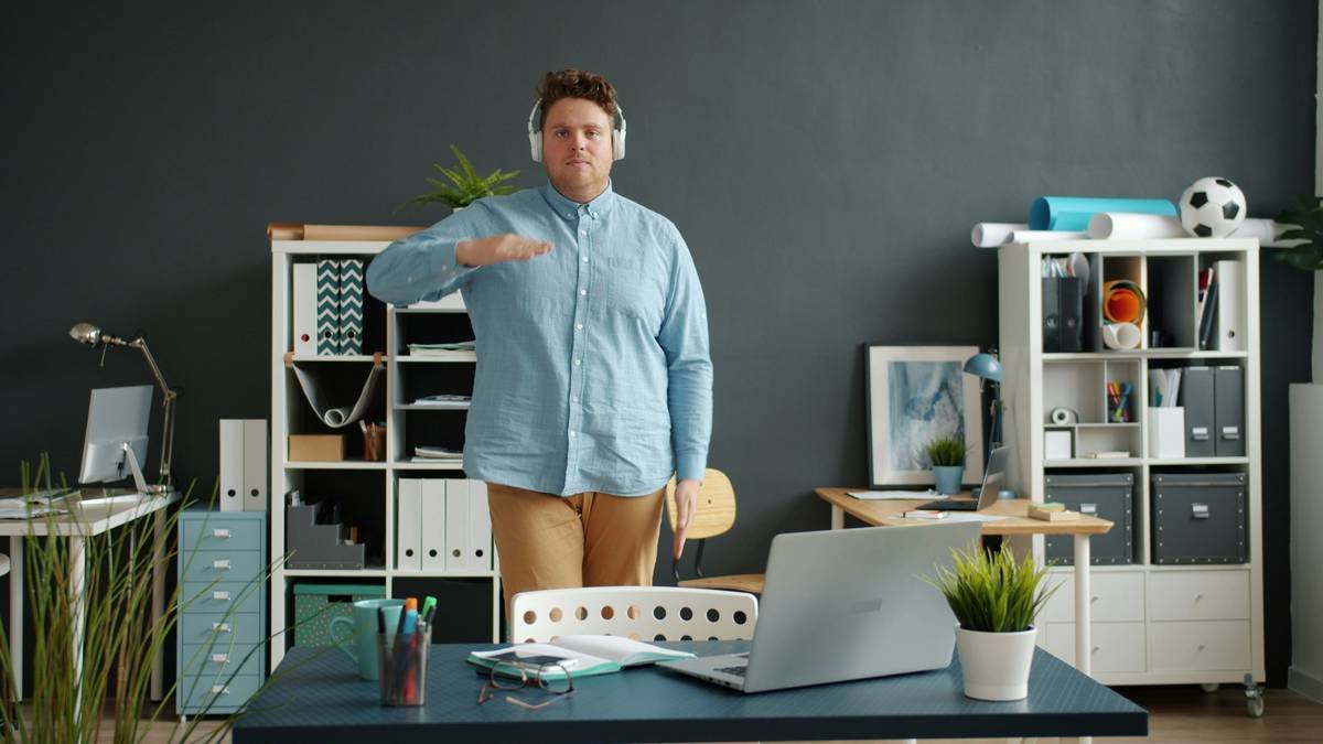 Portrait of handsome funny guy dancing in office room alone wearing wireless headphones looking at camera. Modern devices, people and relaxation concept.